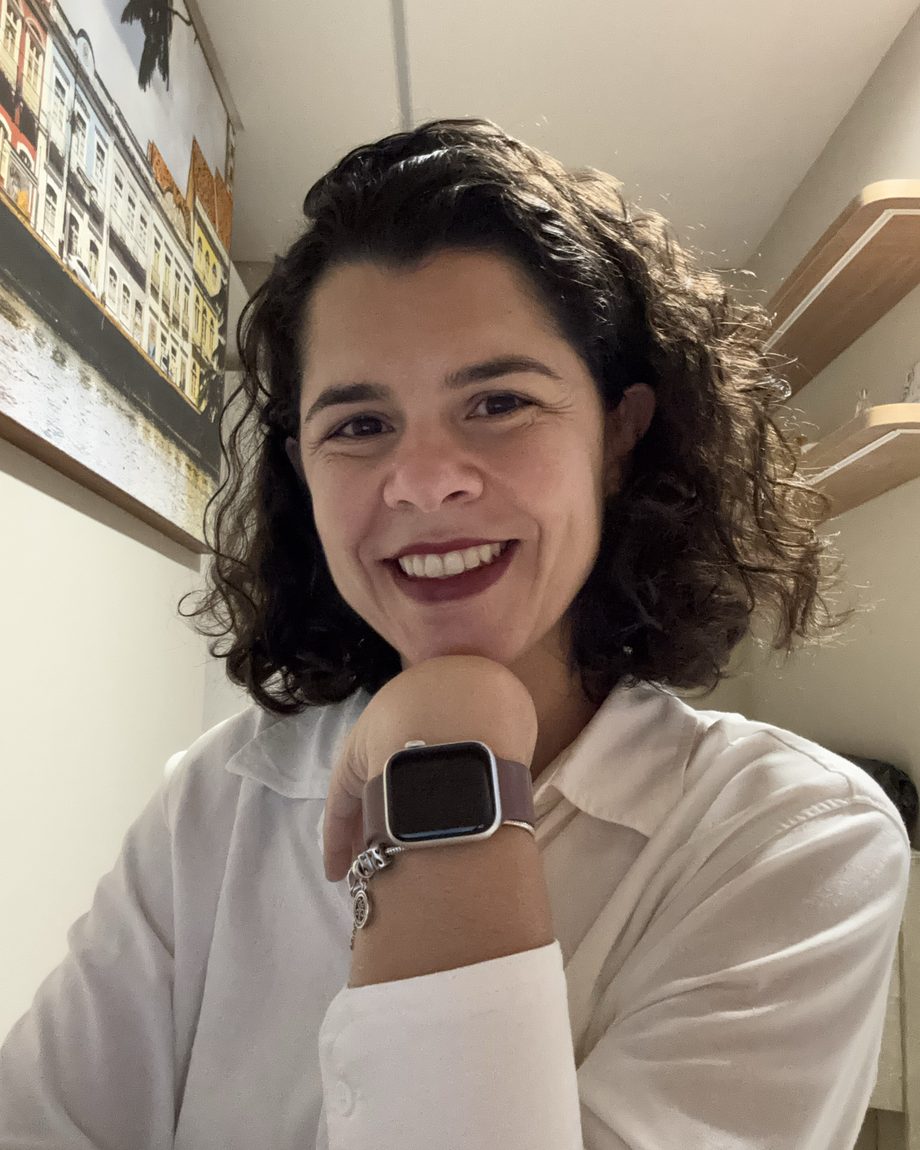 Raquel Tiné, psicóloga clínica em Recife, sorrindo, de camisa clara, apoiando o queixo na mão